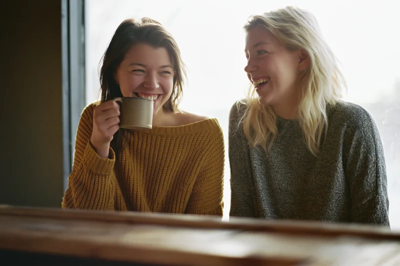 Two women laughing together at a cafe counter holding a coffee mug