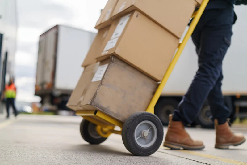 Warehouse worker pushing a yellow hand truck stacked with cardboard boxes at a loading dock