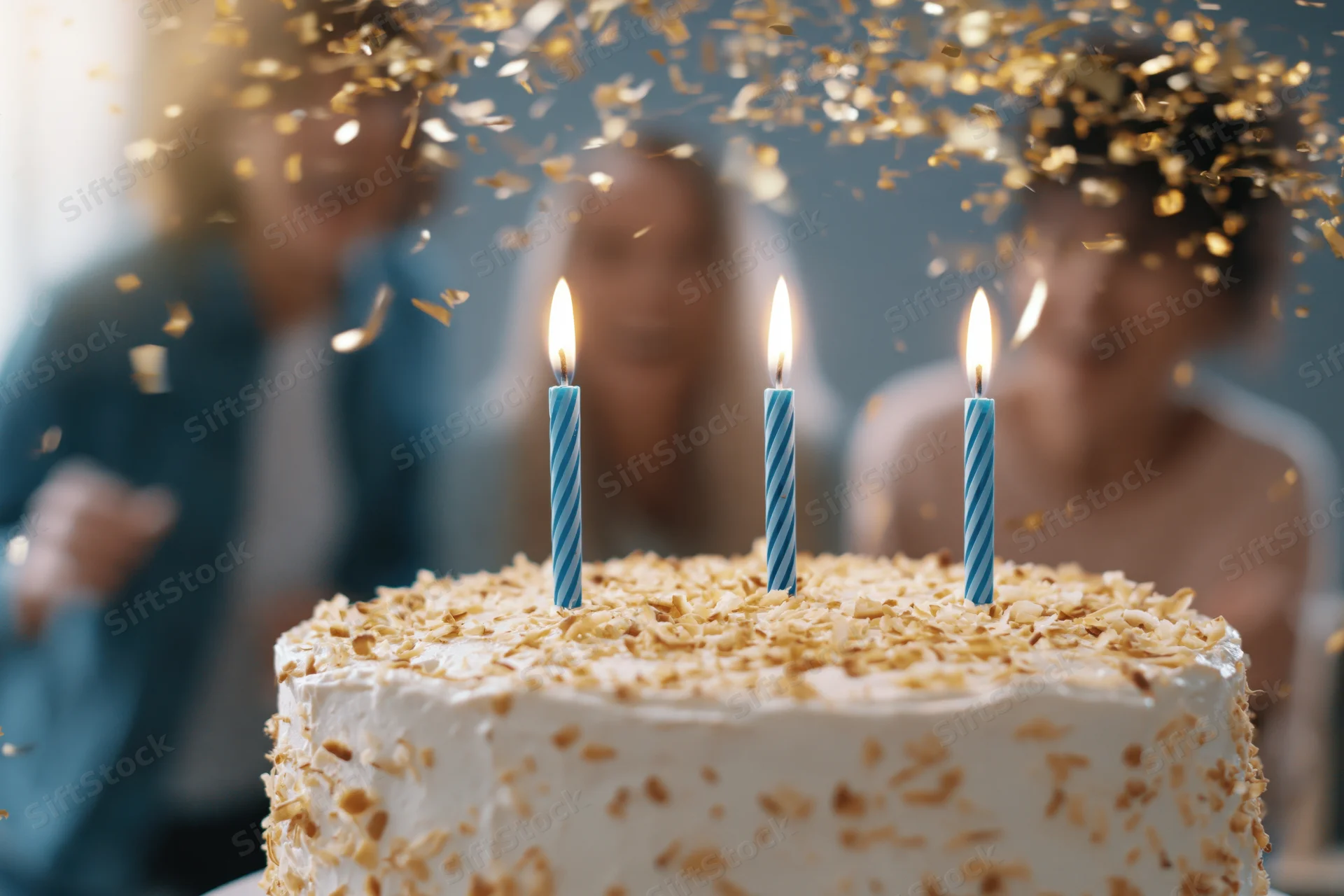 White frosted birthday cake with three lit blue-striped candles and falling gold confetti, with blurred people in the background celebrating
