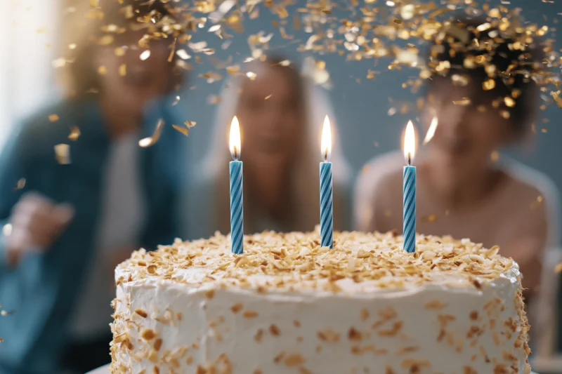 White frosted birthday cake with three lit blue-striped candles and falling gold confetti, with blurred people in the background celebrating