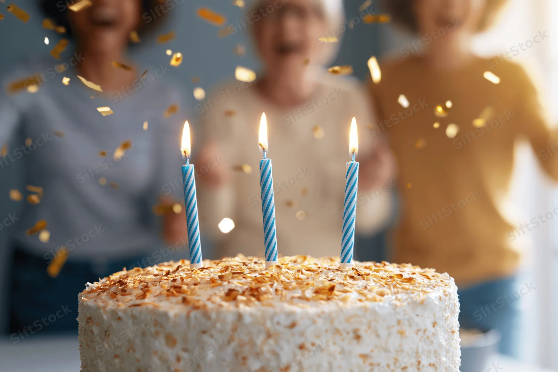 Birthday cake with three lit blue-striped candles, golden confetti falling, people celebrating in background