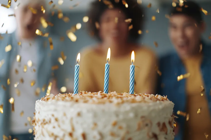 White frosted birthday cake with three lit blue-striped candles in front of smiling young people at a celebration