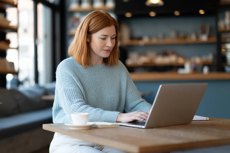 Woman working on laptop at wooden table in cafe with coffee cup nearby