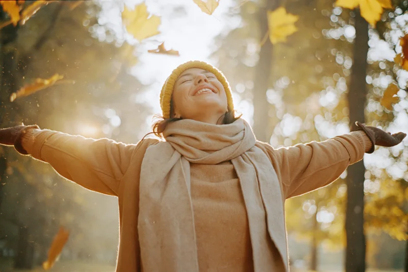 Woman laughing with arms outstretched in autumn forest as leaves fall around her