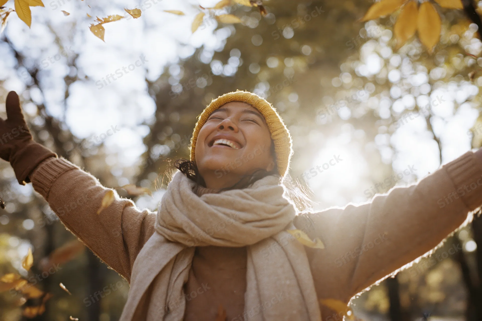 Woman laughing with arms outstretched as autumn leaves fall around her in a sunlit park
