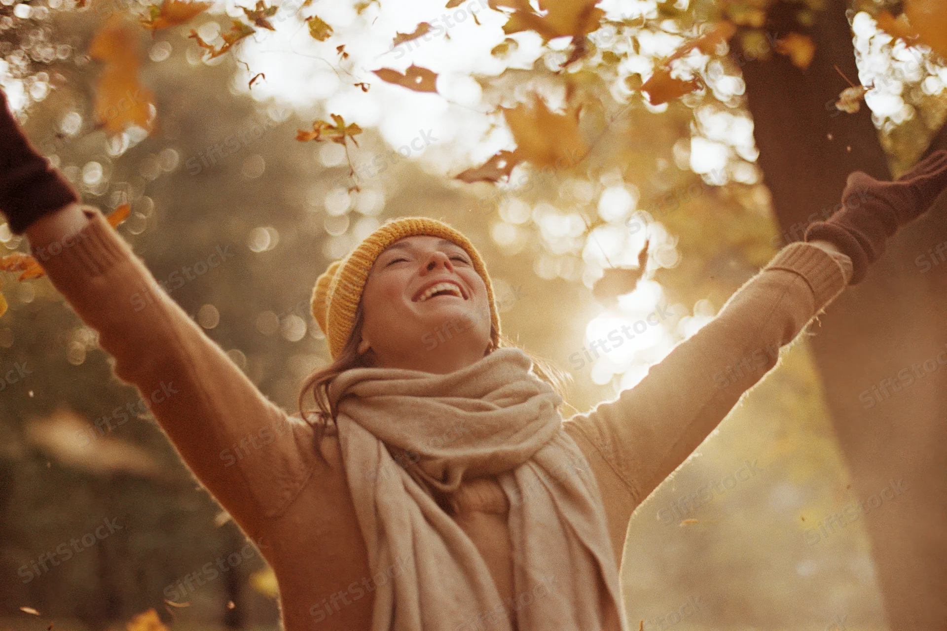 Woman laughing with arms outstretched under falling autumn leaves in a sunlit forest