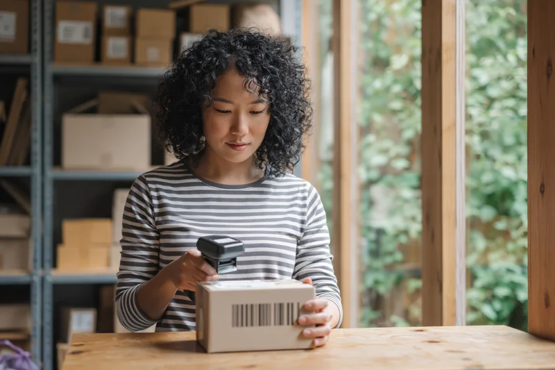 Woman scanning barcode on box at wooden desk in warehouse with shelves and window view