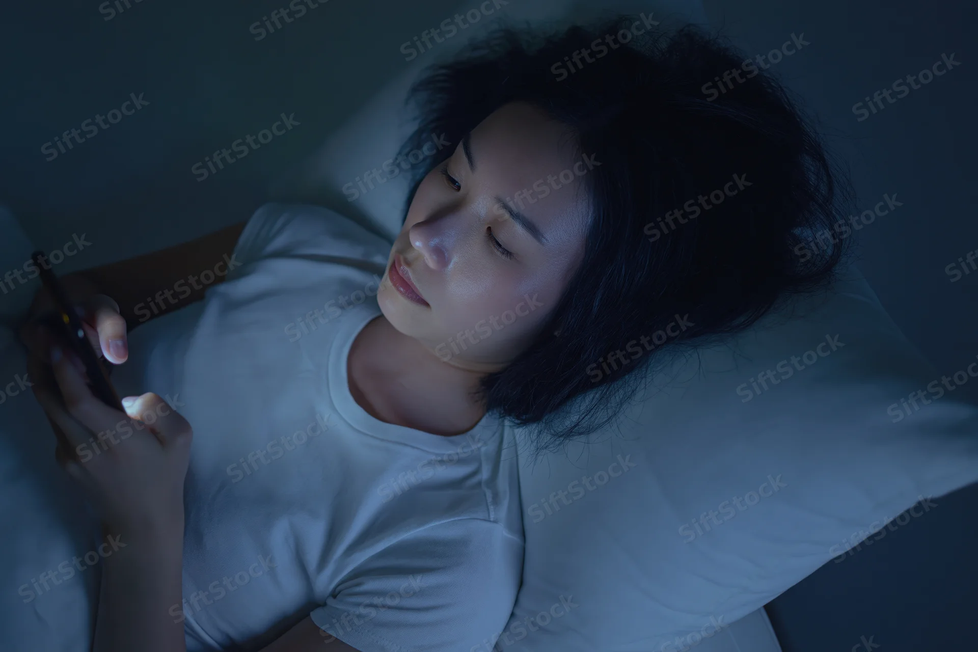 Young asian woman lying in bed at night holding smartphone, illuminated by screen light