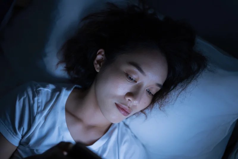 Young asian woman lying in bed at night staring at her smartphone