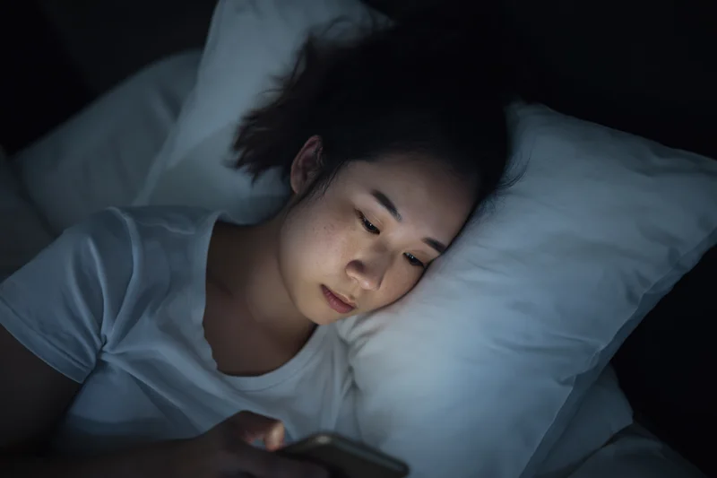 Young asian woman lying in bed at night staring at her smartphone screen