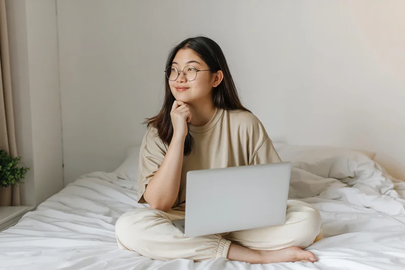 Young asian woman sitting cross-legged on bed with laptop, smiling thoughtfully, in bedroom setting