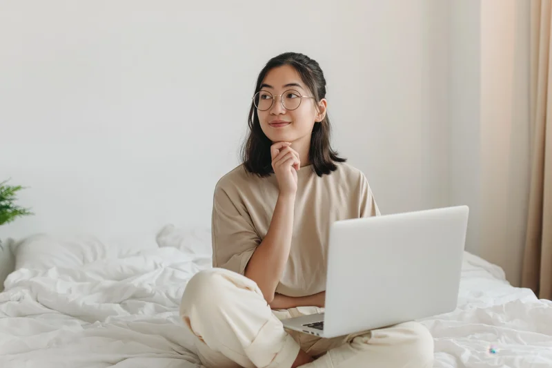 Young asian woman sitting crosslegged on bed with laptop, smiling thoughtfully in home bedroom setting