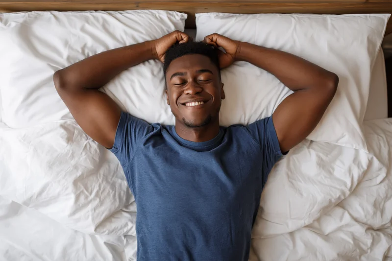 Young black man waking up in bed stretching arms above head with eyes closed and smiling