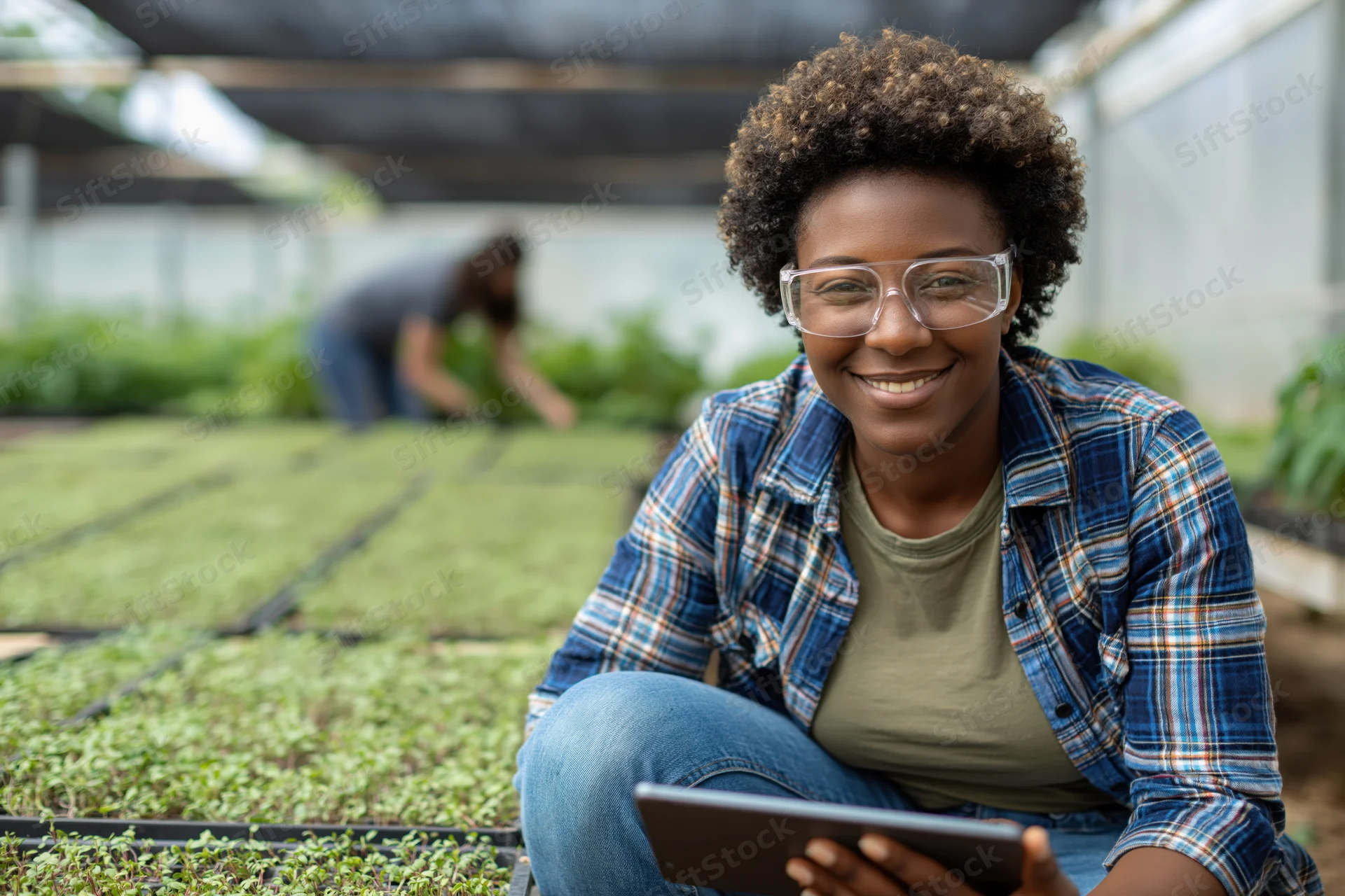 Young black woman smiling while holding tablet beside seedling trays in greenhouse