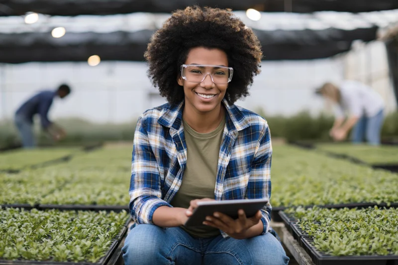 Young black woman smiling while holding tablet beside seedling trays in greenhouse