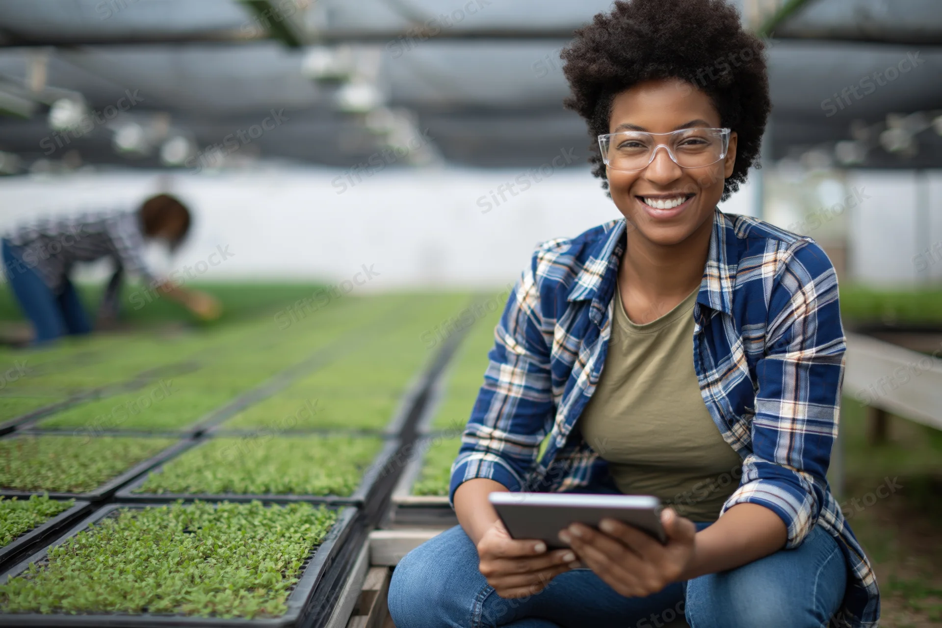 Young black woman crouching with tablet beside seedling trays in greenhouse