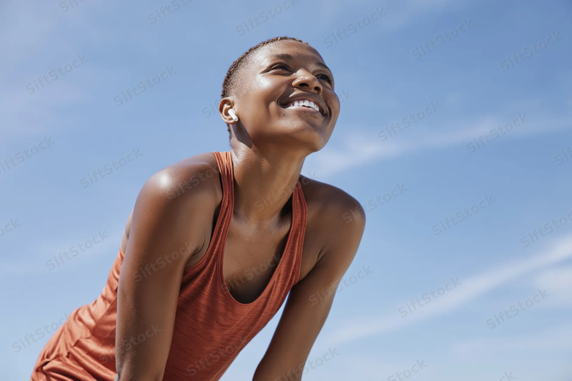 Young black woman smiling during outdoor workout pause with hands on knees against blue sky
