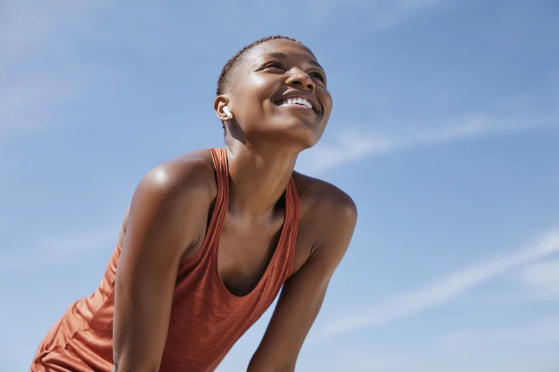 Young black woman smiling during outdoor workout pause with hands on knees against blue sky