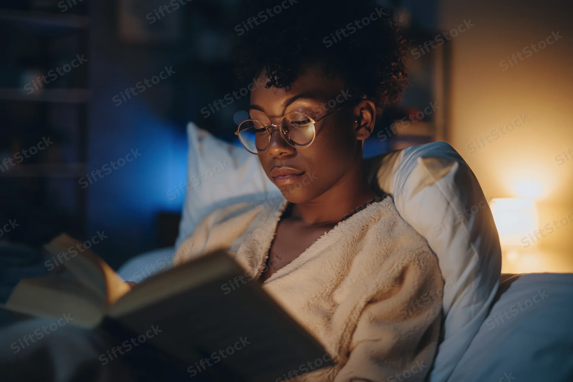 Young black woman reading a book in bed at night, wrapped in a blanket, illuminated by a warm lamp light