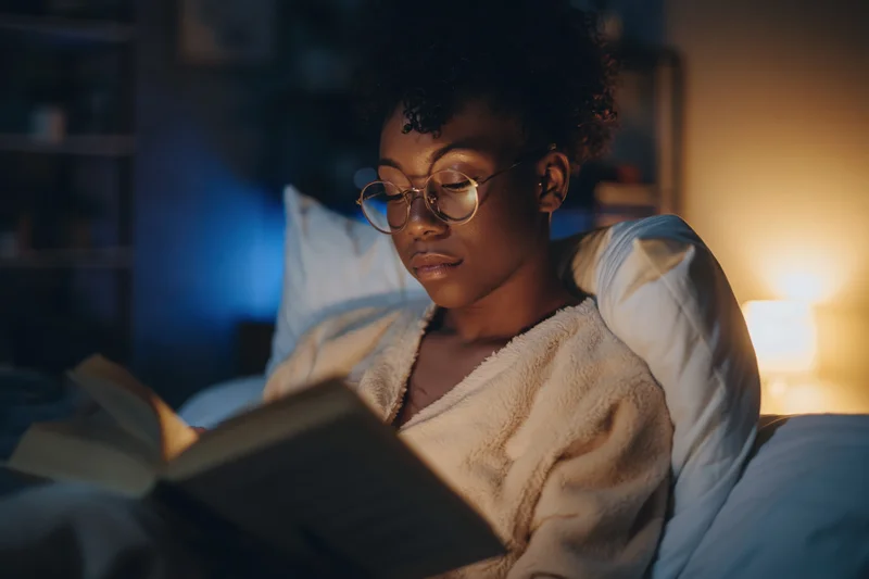 Young black woman reading a book in bed at night, wrapped in a blanket, illuminated by a warm lamp light