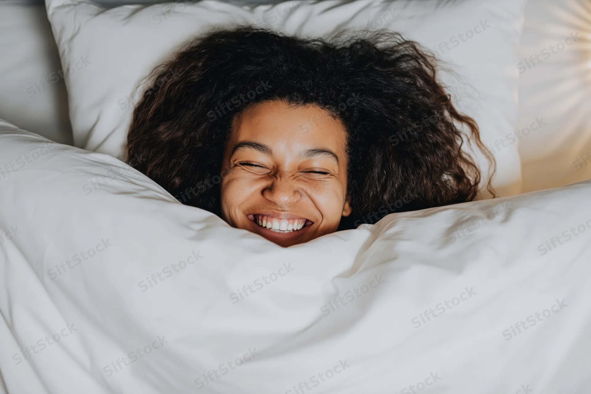 Young black woman laughing joyfully while waking up in bed under white sheets