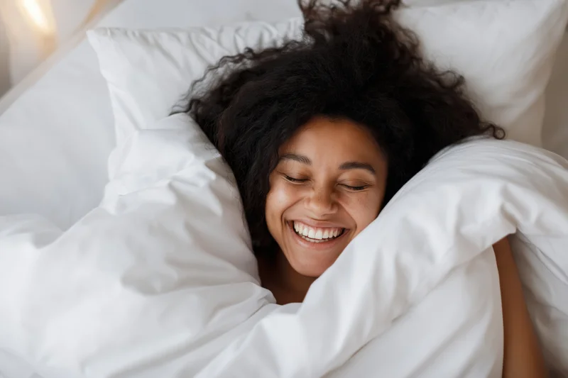 Young black woman laughing joyfully while waking up in bed under white bedding