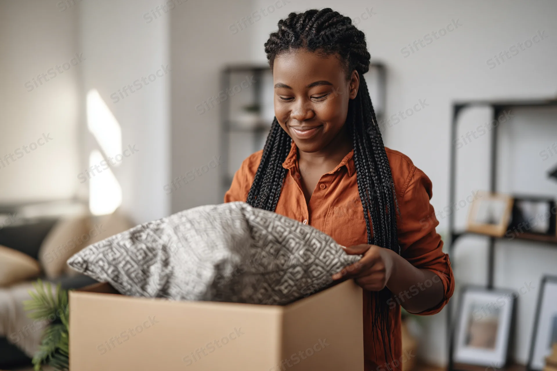 Young black woman unpacking a patterned pillow from a cardboard box in a living room