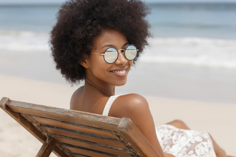 Young black woman smiling while relaxing on wooden lounge chair at beach