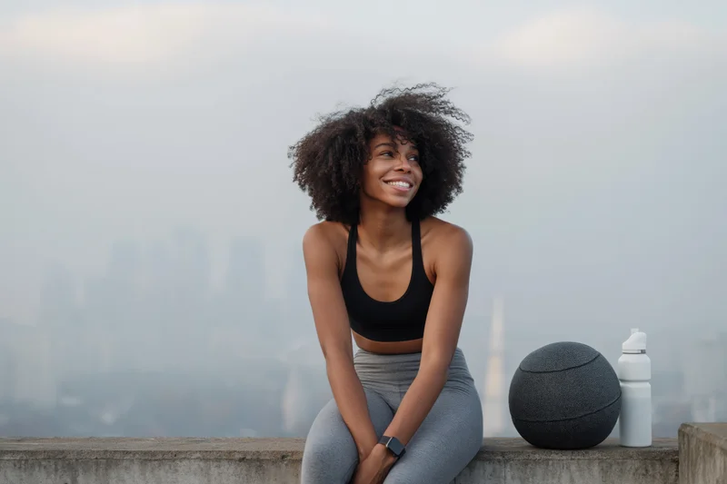 Young black woman smiling while sitting on rooftop ledge with basketball and water bottle, city skyline in background