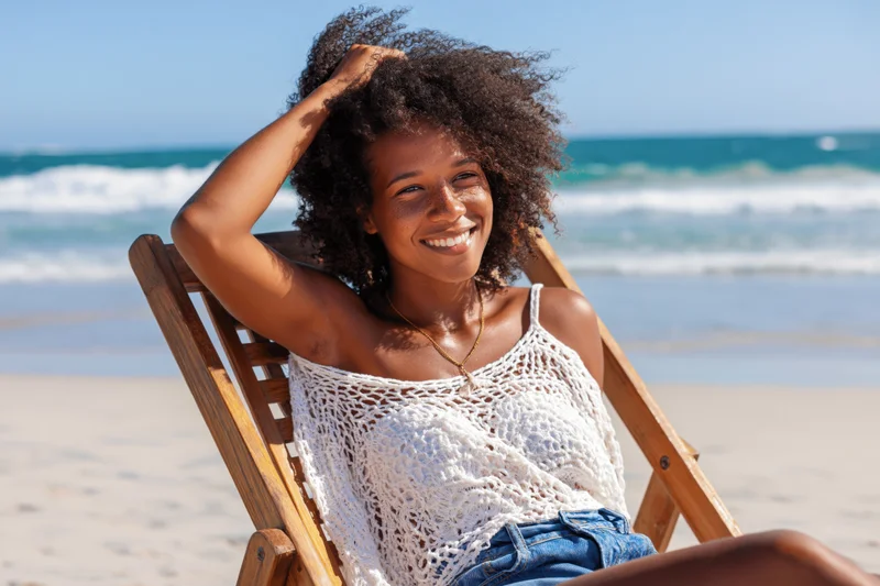 Young black woman smiling while sitting in a wooden deckchair on a sunny beach