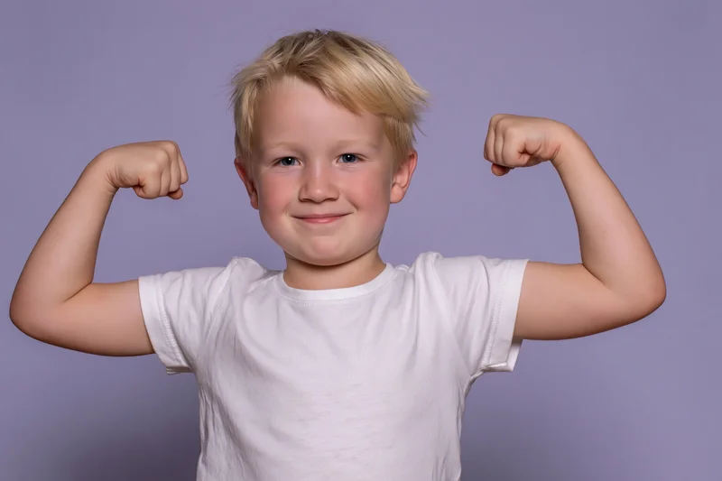 Young blond boy flexing both arms in a strongman pose smiling in front of lavender background