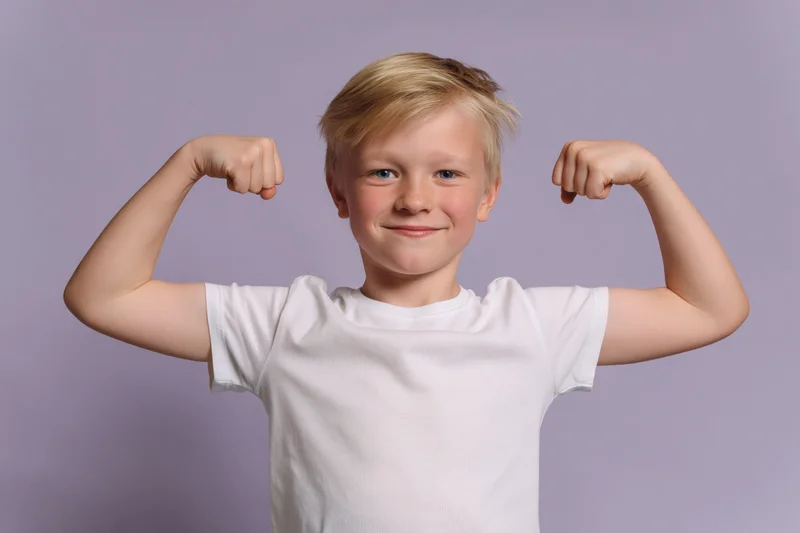 Young blond boy flexing both arms in a strongman pose smiling in front of lavender background