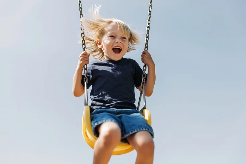 Young blond boy laughing on a yellow swing against a clear blue sky