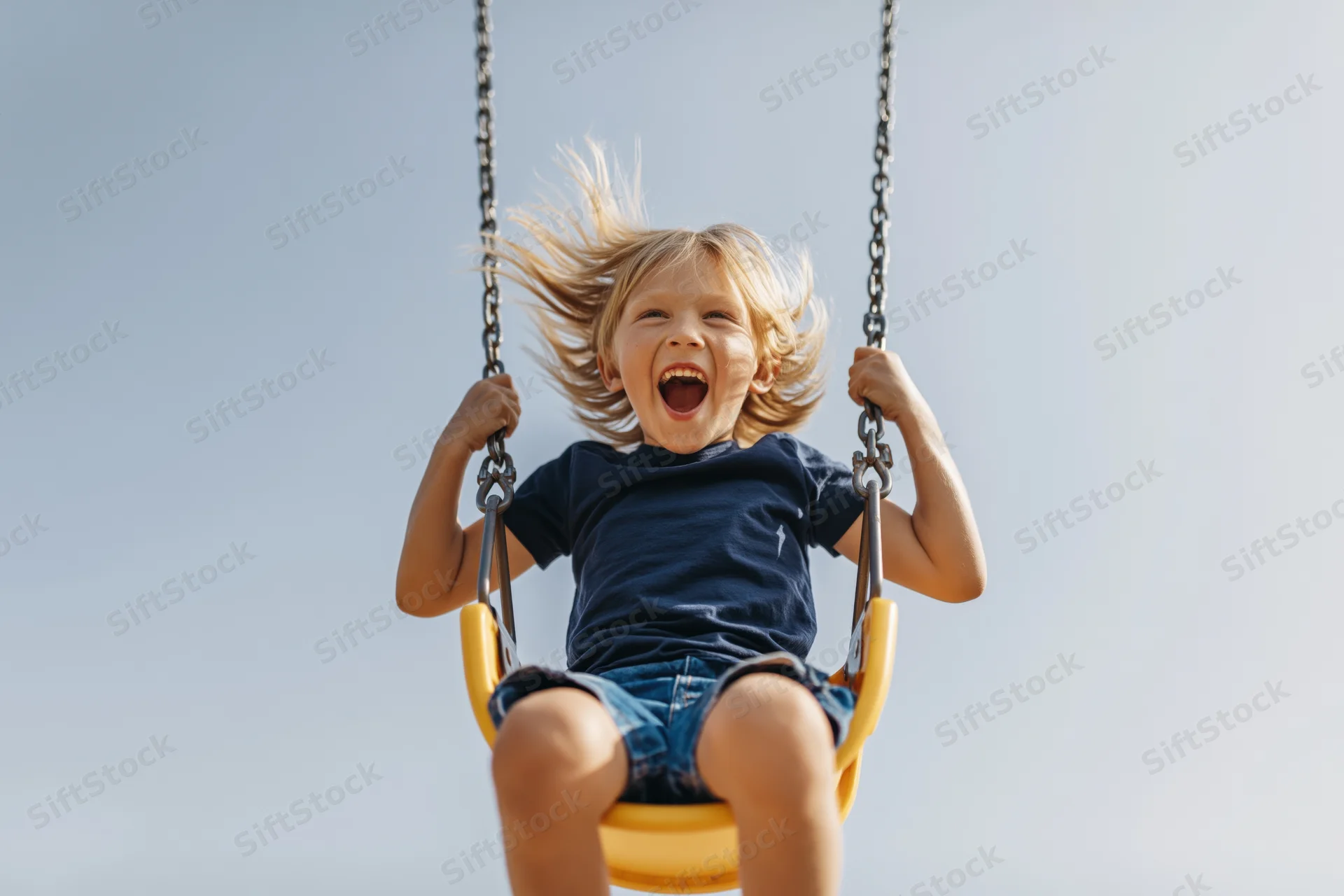 Young blond boy laughing with mouth open while swinging on a yellow playground swing against a clear sky