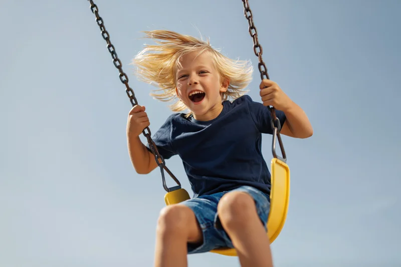 Young blond boy laughing on a yellow swing against a clear blue sky