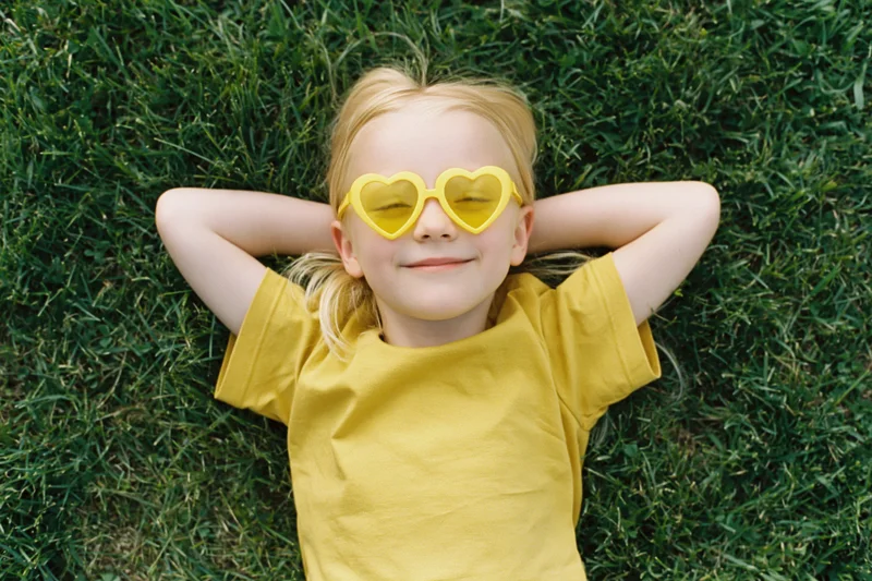 Young blond girl lying on green grass with hands behind head wearing yellow heart-shaped sunglasses and smiling