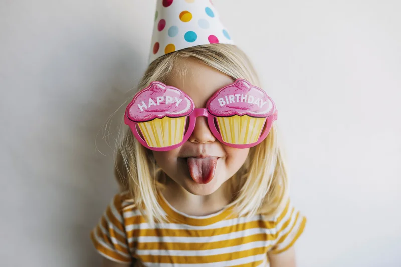 Young blonde child sticking tongue out while wearing cupcake-shaped birthday glasses and a polka-dot party hat indoors