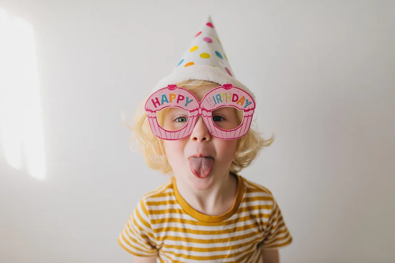 Young blonde child sticking tongue out while wearing birthday hat and cupcake glasses indoors