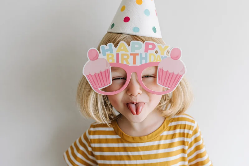 Young blonde girl sticking tongue out while wearing birthday party hat and cupcake glasses against white background