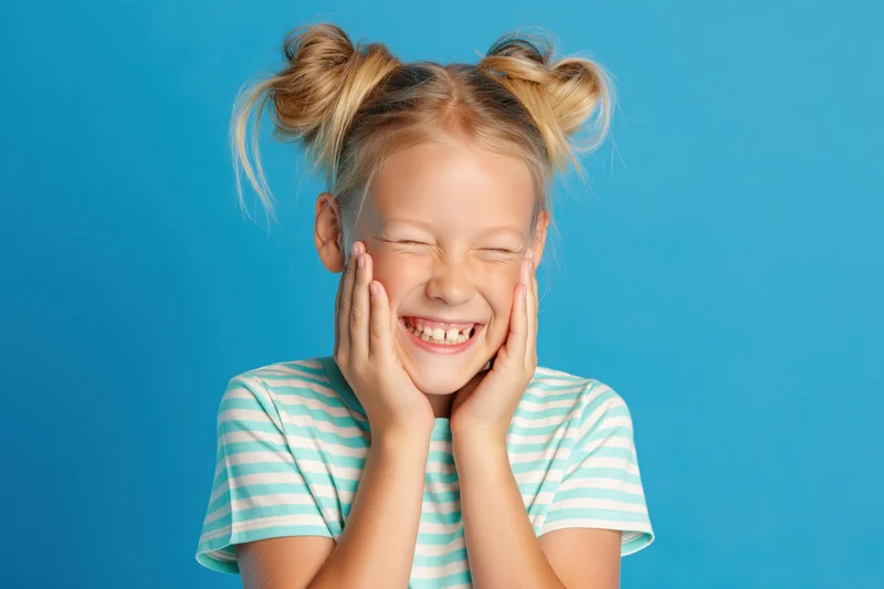 Young blonde girl laughing with hands on cheeks against blue background