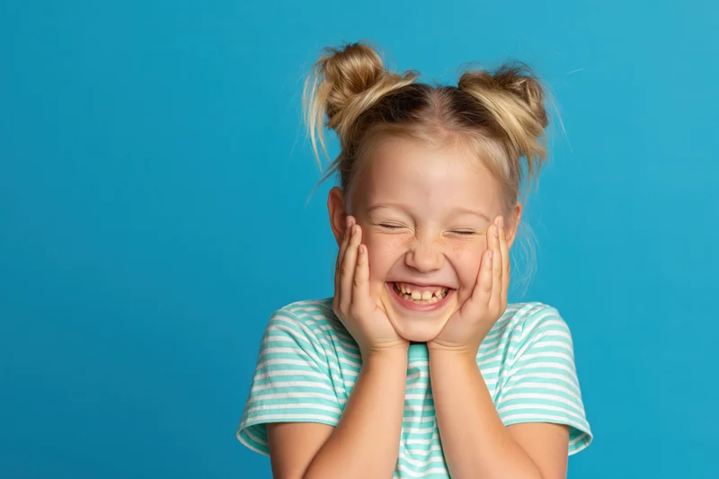 Young blonde girl laughing with hands on cheeks against blue background