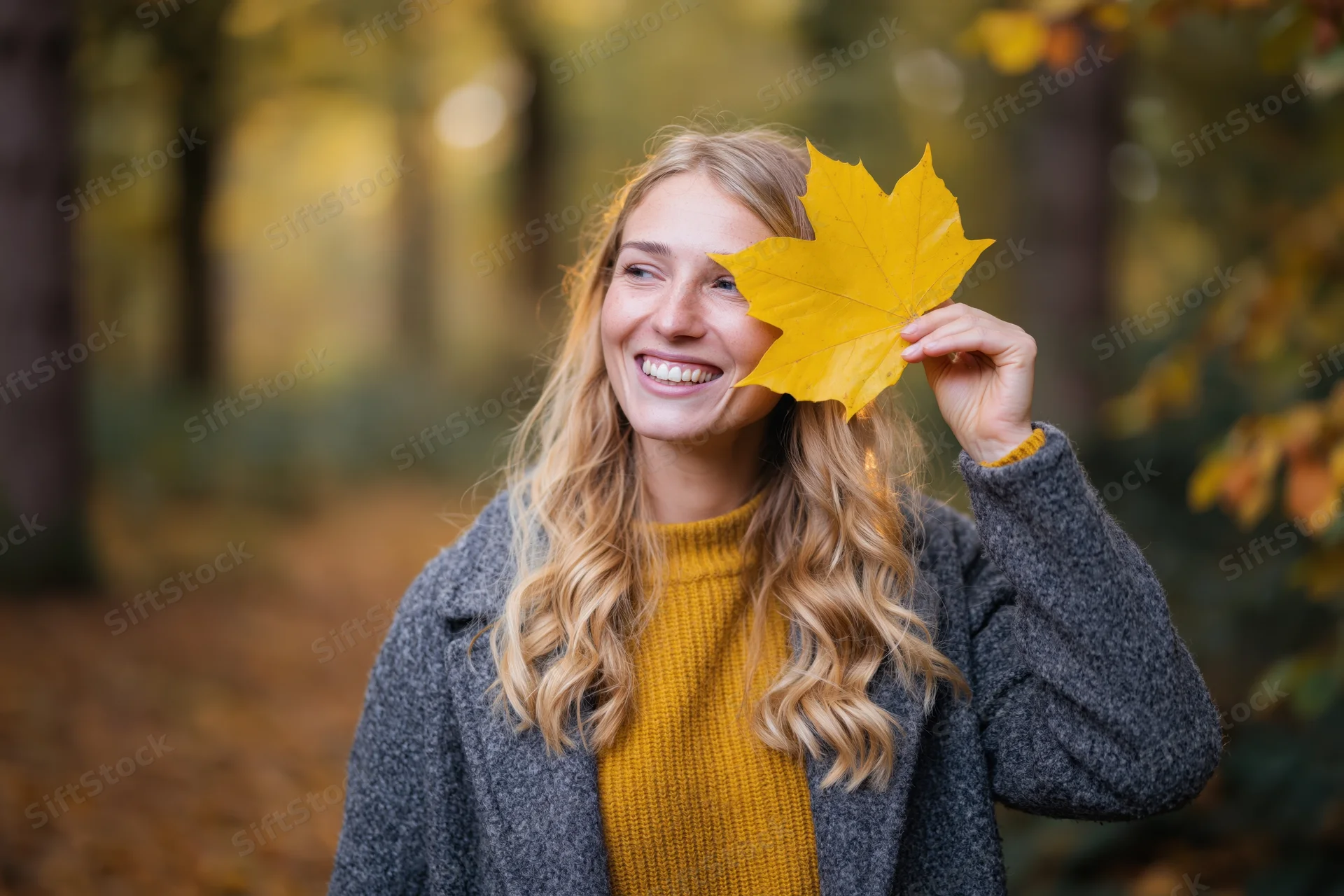 Smiling blonde woman holding yellow maple leaf in autumn forest