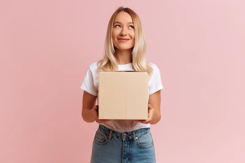Young blonde woman holding a cardboard box and looking up against a pink background