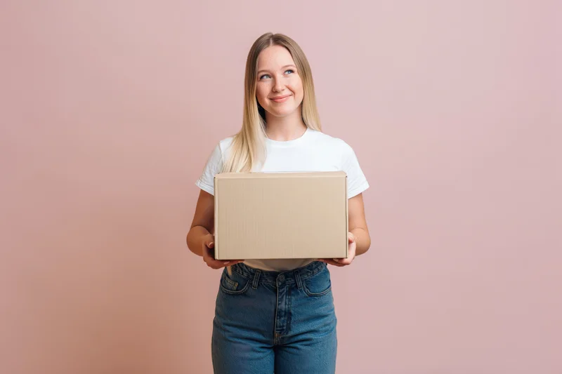 Young blonde woman holding a cardboard box against a pink studio background