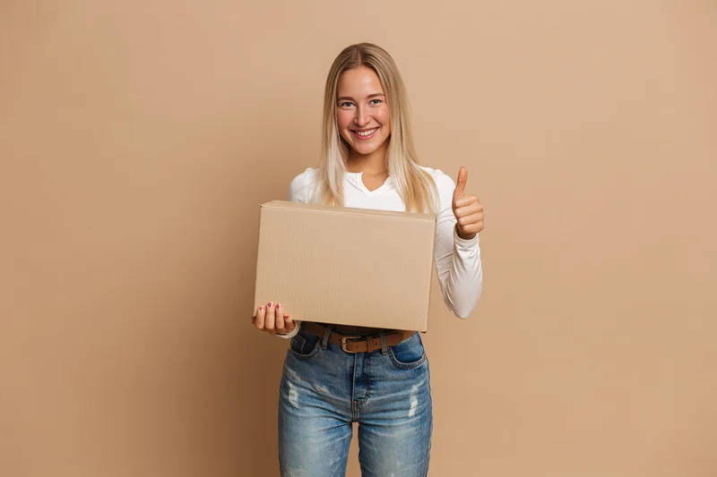 Young blonde woman holding cardboard box and giving thumbs up against plain background