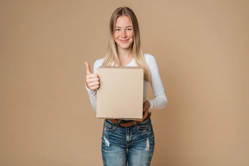 Young blonde woman holding a cardboard box and giving thumbs up against beige background