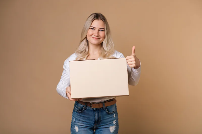 Young blonde woman holding a plain cardboard box and giving thumbs up against a beige background