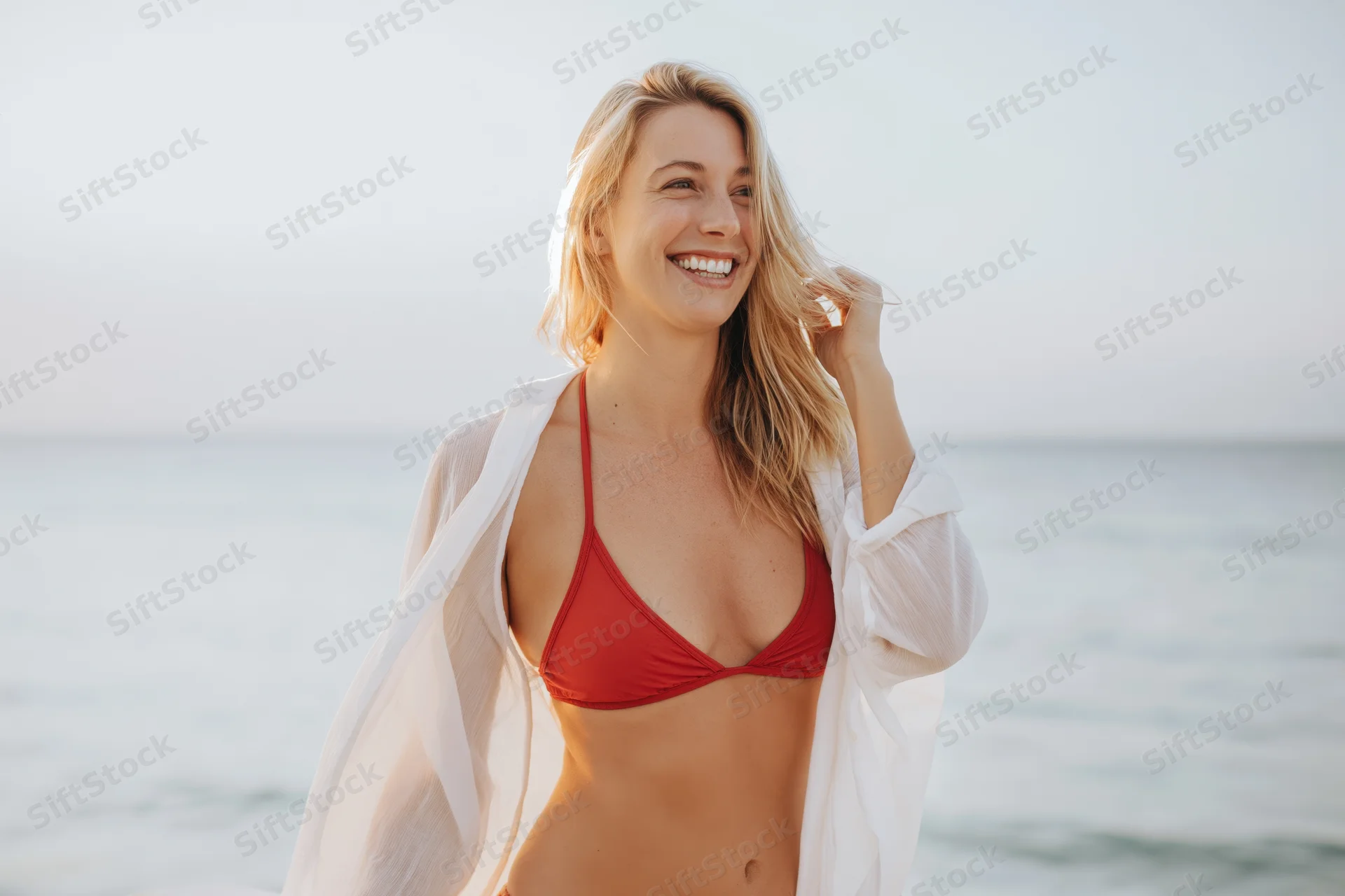 Smiling blonde woman in red bikini with white coverup standing on beach by ocean
