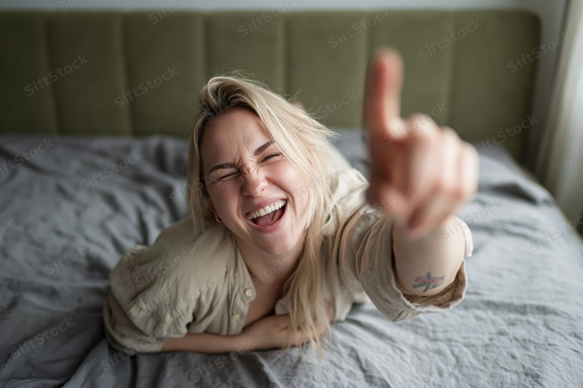 Young blonde woman laughing and pointing finger while lying on stomach on bed
