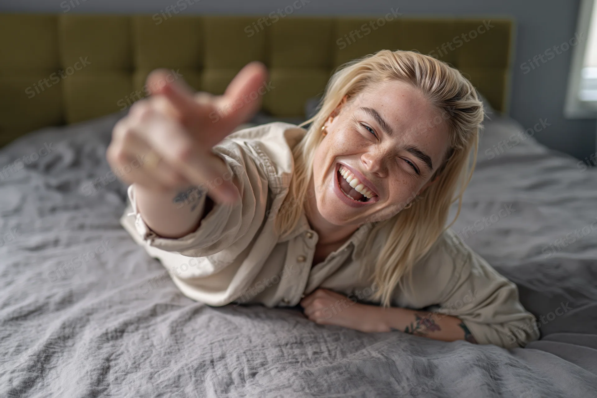 Young blonde woman laughing and pointing from bed in bedroom